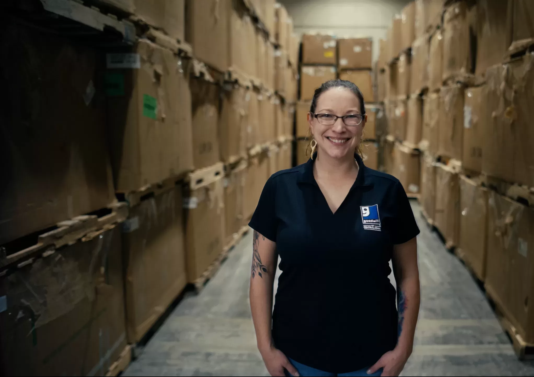 woman with glasses and goodwill polo stands in a room full of cardboard boxes