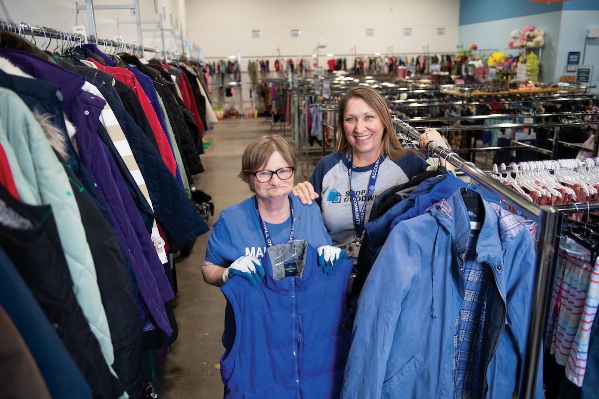 two women stand together in a goodwill store, holding up merchandise and smiling